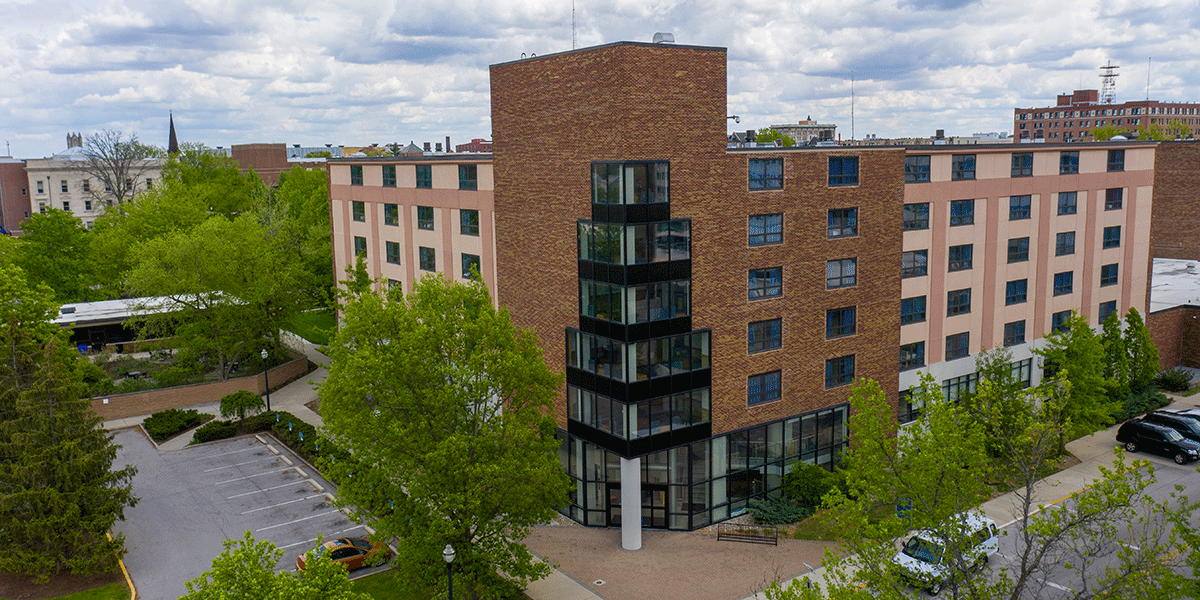 A brick building with glass windows. A parking lot is to the left and green trees are in the front. 
