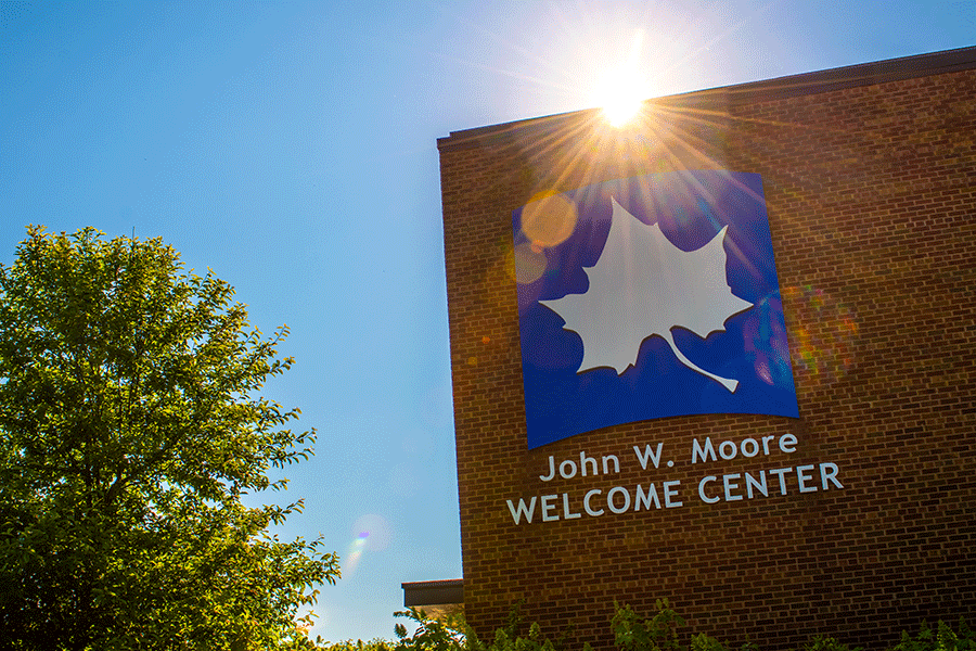 The sun is partially visible over the red brick wall of the John W. Moore Welcome Center. Rays of light shine over top the large white Sycamore leaf on the blue background that appears on the side of the building with the name underneath. A green tree is also visible to the right against the blue sky. 