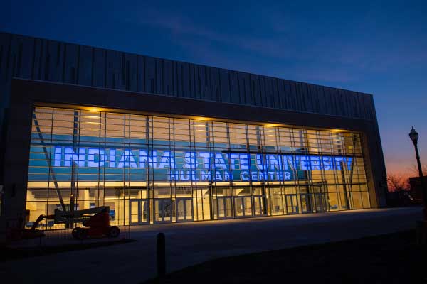 An exterior photo of Indiana State University’s Hulman Center arena in the evening. The building has clear glass windows in the front with “Indiana State University Hulman Center” in blue lettering, illuminated. A light post is visible to the right against the darkening sky.