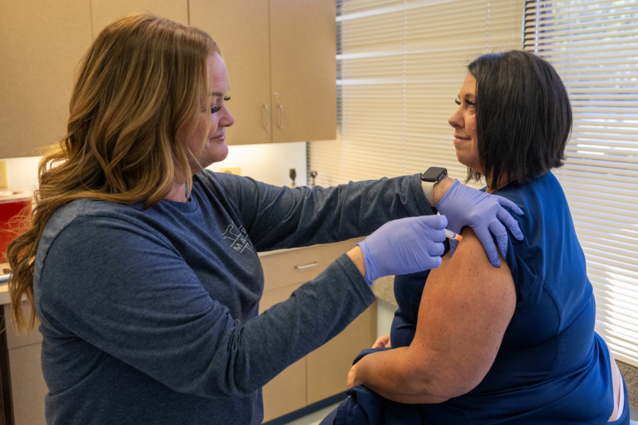 A smiling woman with dark blonde hair wearing a gray sweater and blue lab gloves administers an injection to a woman with short, dark brown hair wearing a blue shirt with a sleeve rolled up. They are standing in an exam room in front of a window with closed blinds. White cabinets are visible behind them.