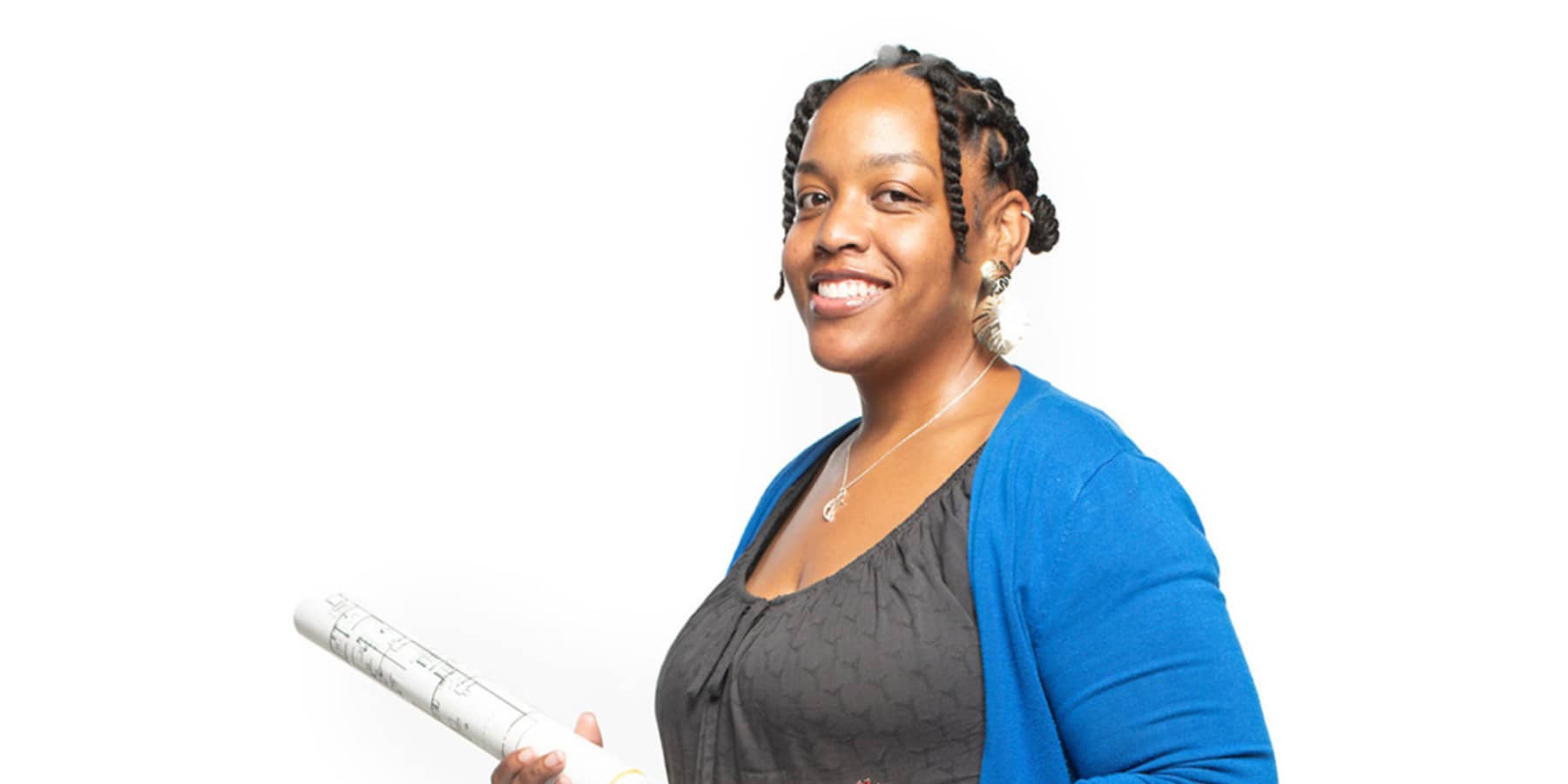 Professor of Interior Architectural Design Azizi Arrington-Slocum, a smiling Black woman with black braided hair, wearing a grey shirt with a blue jacket, and holding a set of rolled-up architectural designs.