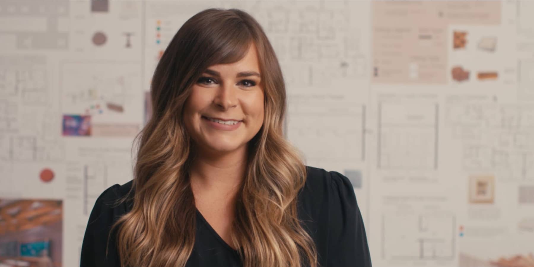 A woman with wavy brown hair poses with interior design sketches and designs behind her. She wears a black dress shirt.