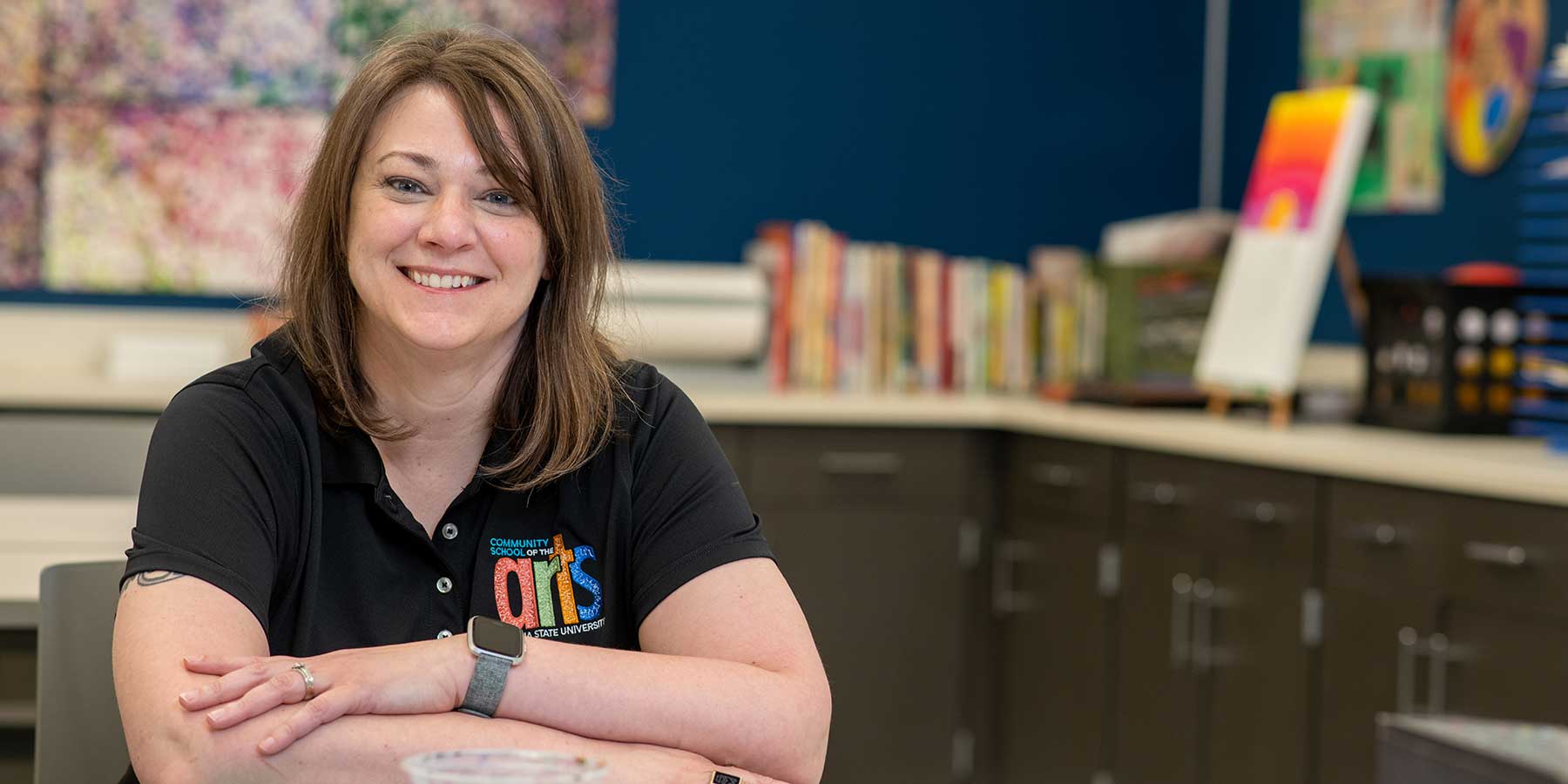 A white woman with shoulder-length brown hair sits, smiling at the camera. She wears a black T-shirt with a colorful patch reading Community School of the Arts, Indiana State University. Cabinets and counters are visible behind her with art supplies and art projects