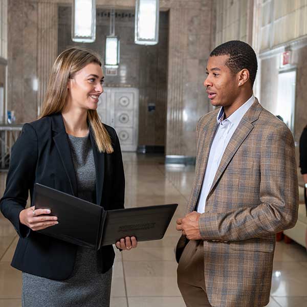 Two individuals standing in a hallway. On the left is a white woman with straight dark blonde hair. She wears a grey dress and a black jacket. She holds an open black folder. On the right is a Black man with short brown hair. He wears a tan-and-blue checkered suit and a white dress shirt