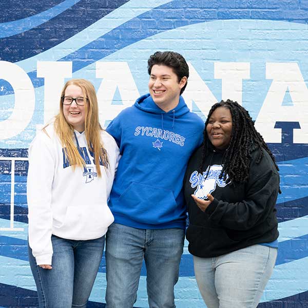 Three students standing in front of a blue-and-white painted Indiana State mural. On the far left is a white female student with straight blonde hair. She wears a white Indiana State sweatshirt, black glasses, and blue jeans. In the middle is a white male student with black hair. He wears a blue Sycamores sweatshirt and blue jeans. On the right is a Black female student with black dreadlocks. She wears a black Sycamores sweatshirt and blue jeans.