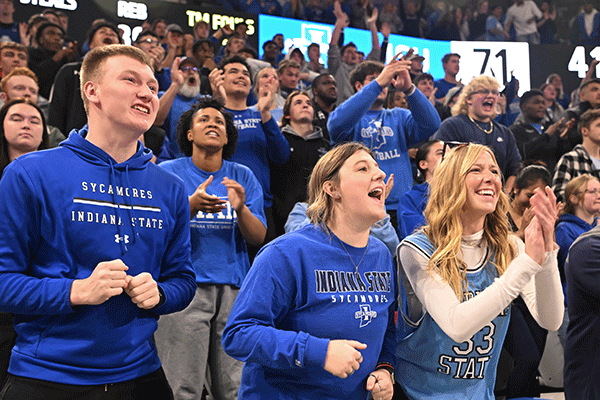 A diverse group of smiling Sycamore fans, most wearing various shades of blue Indiana State clothing, cheer and clap at a home Indiana State basketball game in the Hulman Center. Digital signage and a scoreboard are just visible behind them.