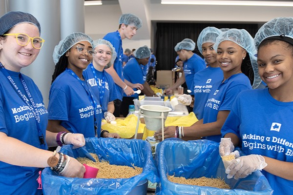 A diverse group of students smiles at the camera while packing food at a volunteer event. They are all wearing hairnets and shirts that say “Making a Difference Begins With Blue.” Other students are visible working in the background.