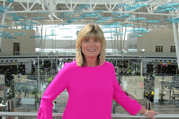 A woman with blonde hair and dressed in a pink blouse smiles directly at the camera. She is standing in an elevated position in front of an airport terminal.