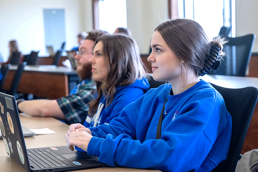 Three students – two women with brown hair in blue sweatshirts, and a man with facial hair and a checkered shirt – sit in front of laptops as they look toward the front of a classroom. The woman nearest the camera is smiling softly.