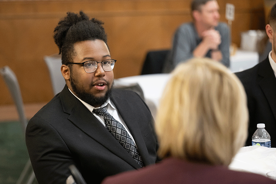 A man in a black business suit with white shirt and grey checkered tie is seated at a table in a conference room. He is talking with another person whose face is not visible.