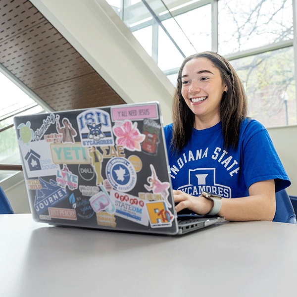 A female student wearing a blue T-shirt types on a laptop which is covered with stickers