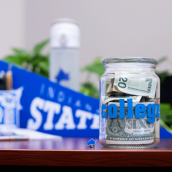 A clear glass jar with ‘COLLEGE’ printed on it in blue letters sits on a desk. It is filed with bills of various denominations. A folded $20 is visible, as is the back of a $1 bill. A blue Indiana State pennant is visible but out of focus in the background, along with a white and gray thermos with a blue Sycamore leaf on it, a sheet of stickers, and a few other items. Greenery and a white wall are visible in the background.