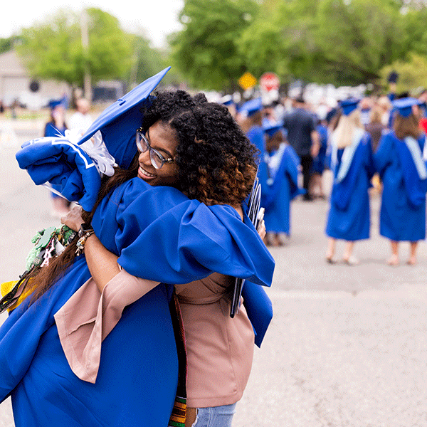 A smiling young woman of color with glasses and dark, curly hair hugs an Indiana State graduate in blue cap and gown outdoors on a spring day. Other graduates and guests are visible in the background, as are many green trees, traffic signs, and buildings in the distance.