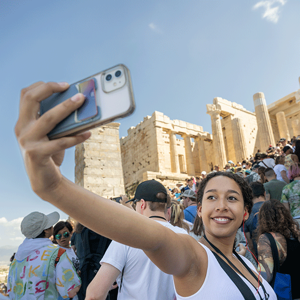 A smiling young woman of color wearing a white tank top takes a selfie with the Greek Parthenon in the background. The hand holding her phone is pictured in the foreground, and the Parthenon is visible at a high angle on a hill behind her amidst crowds of people.