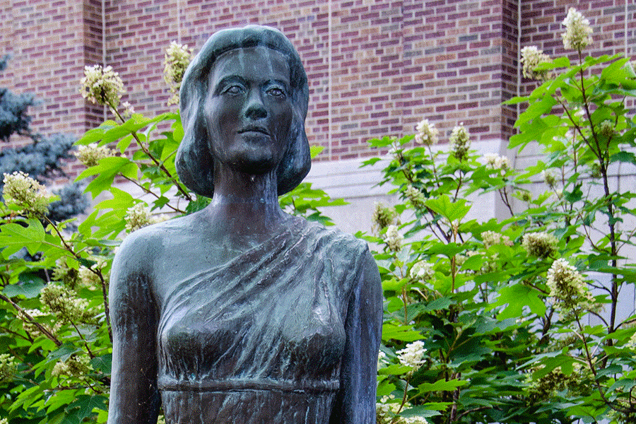 A concrete statue of a woman wearing an off-the-shoulder toga. The statue depicts shoulder-length hair on the woman. Behind the statue, green plants are visible, and a brick building can be seen in the background.