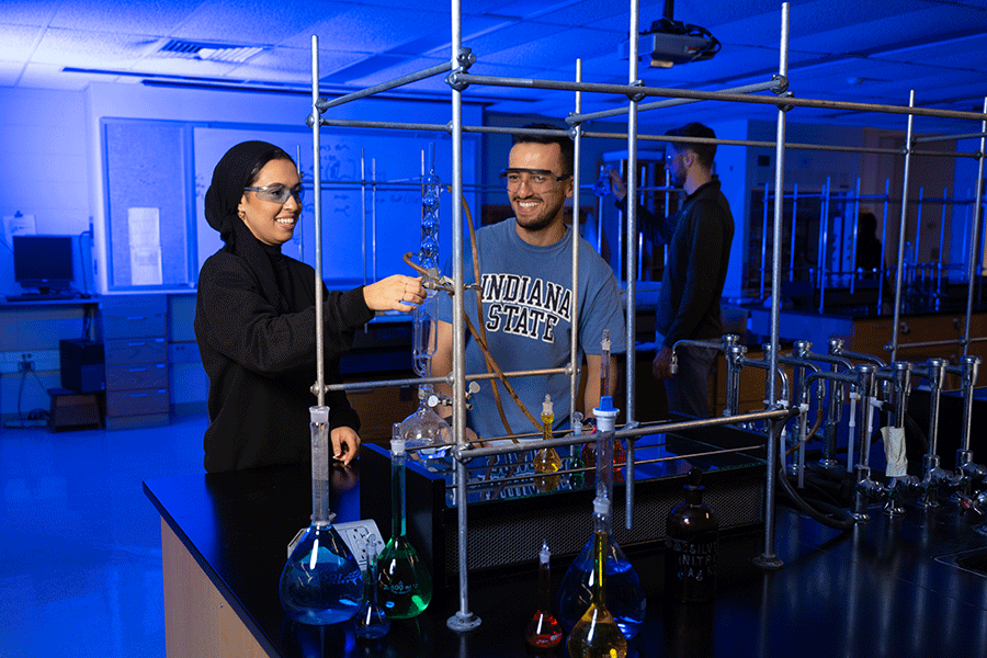 Three students of different ethnicities stand in a blue-lit lab. On the left is a female student wearing a black outfit and black hijab. To her right is a male student with short brown hair. He wears a blue Indiana State T-shirt. Behind them is a male student in a black jacket. The students are wearing safety glasses, and the female student is pointing to a long, tall test tube.  