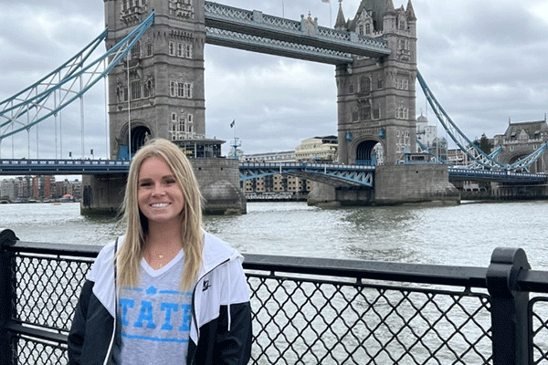 A female student with blonde hair poses in front of the London Bridge, a bridge with two stone towers. Water is behind her. She wears a grey-and-blue STATE T-shirt and a black-and-white jacket.