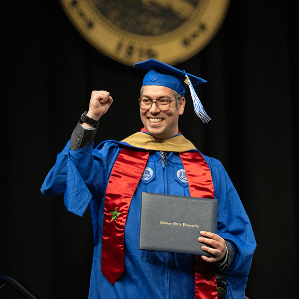 A student in a cap and gown walks across the stand holding a diploma case