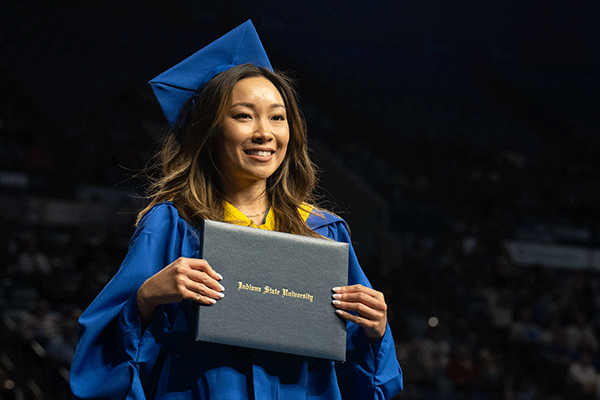 A student in a cap and gown holds a diploma case on stage