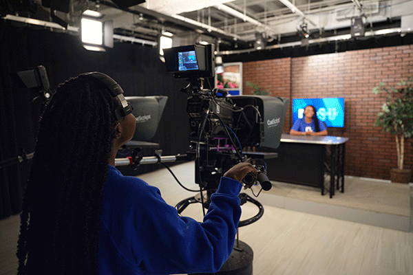 Inside a media newsroom, a Black student with long black dreadlocks stands behind a black camera and teleprompter, wearing a blue sweatshirt. On the set is a Black student wearing a blue T-shirt, blurred in the photo. A desk and television are visible on the set. A brick wall is in the background. Ceiling lights are also visible in the newsroom. 