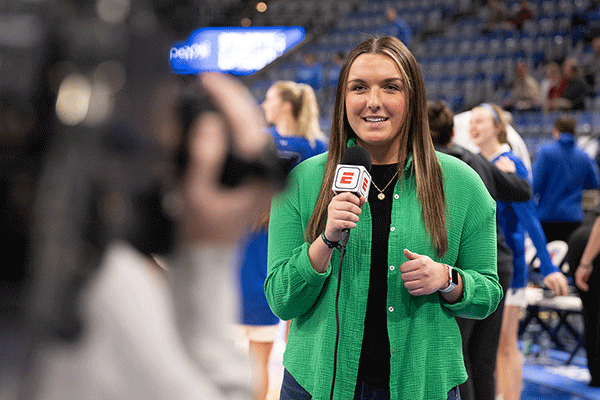 A white female student with long brown hair is at a basketball game inside the Hulman Center. She wears a green sweater vest with a black undershirt. She holds a microphone with a red “E” on a white square on the microphone. She is looking toward a cameraperson holding a black camera. Cheerleaders in blue uniforms are visible behind her. The arena’s bleachers are also visible in the background. 