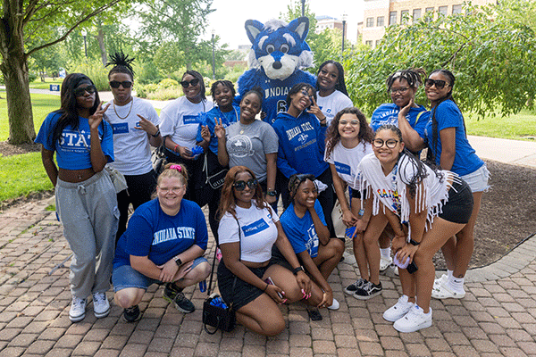 A diverse group of 14 smiling female students pose in front of Sycamore Sam on a campus walkway at Indiana State. Everyone is wearing blue or white shirts. Those in the back row stand beside Sam while those in the front row kneel in front.