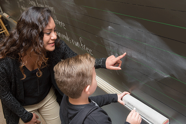 A white female student with long curly brown hair is kneeling at a chalkboard with a young boy. She wears a black long-sleeved shirt and khaki pants. The boy has short brown hair and wears a black hoodie. The female student points to the chalkboard.