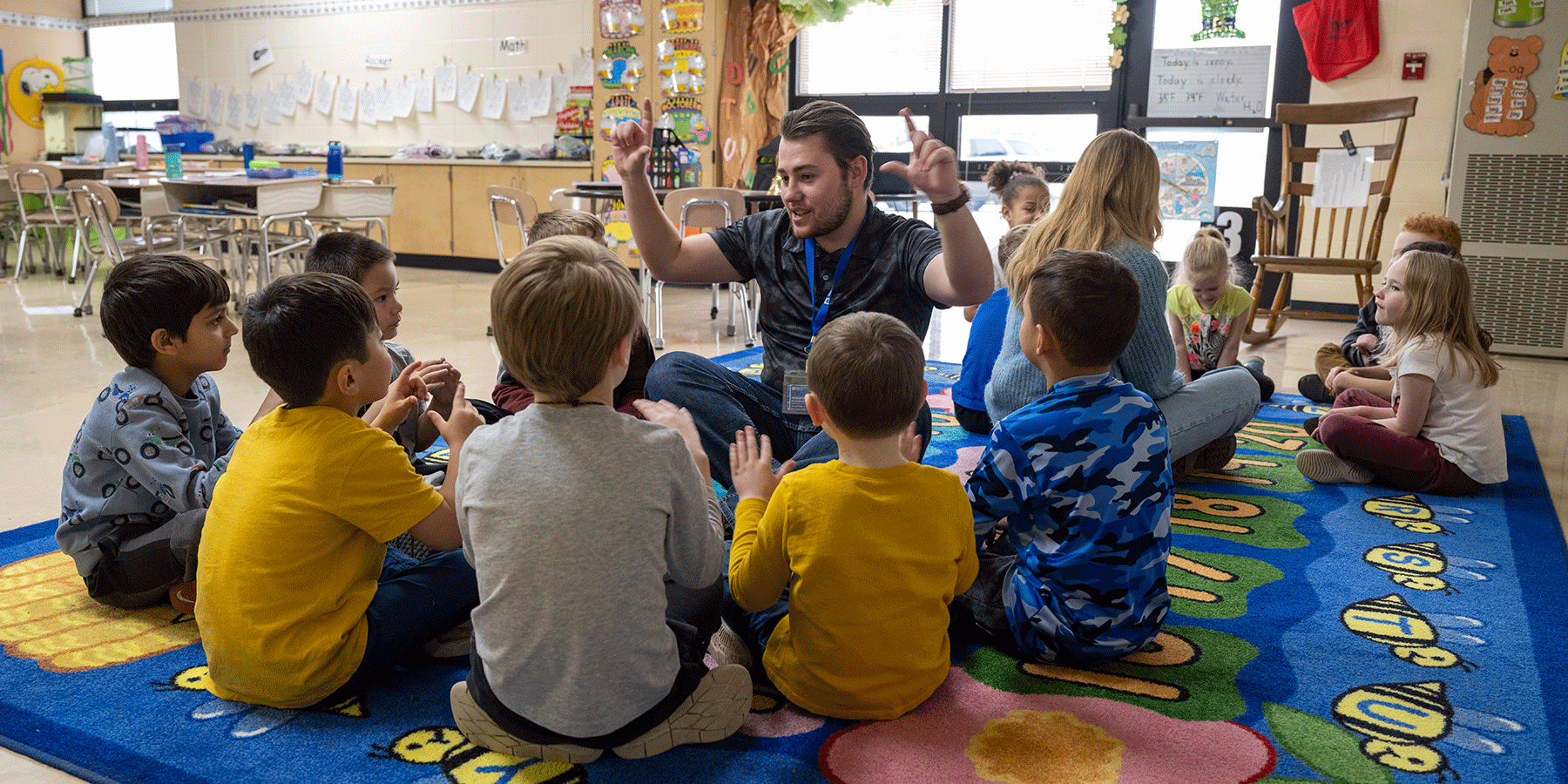 A classroom with 13 young children of mixed races, wearing a variety of yellow, blue, and grey clothing. The children are divided into two groups, sitting in circles on a blue, green, and yellow-colored floor rug. In the left group, a white male sits with the students. He has short brown hair with a matching beard. He wears a black T-shirt and blue jeans. He is holding up both hands as he looks at the students. In the right group, a white female sits with the students.