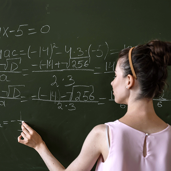 A white female student stands at a chalkboard. She has brown hair pulled back into a bun. She wears an orange headband and a light pink dress. She holds a piece of chalk in her left hand, writing on the chalkboard. Numbers and mathematical equations are written on the chalkboard.