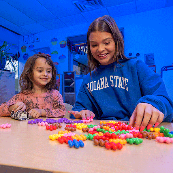 A white female student with shoulder-length brown hair sits at a table with a young girl with shoulder-length brown hair. The female student wears a blue Indiana State sweatshirt. The young girl wears a pink long-sleeved shirt. Multi-colored beads are on the table. The room is blue-lit in the background.