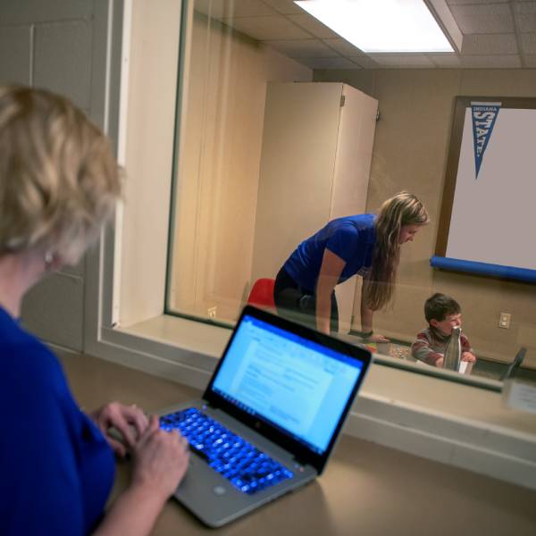 A white female with short blond hair stands in a room with a laptop on a counter in front of her. She wears a blue dress. A glass window separates her from a classroom, where she is observing a white female student with long blond hair, wearing a blue T-shirt and black pants. The student is leaning down to a toddler boy with short brown hair. A filing cabinet and a whiteboard are in the background.