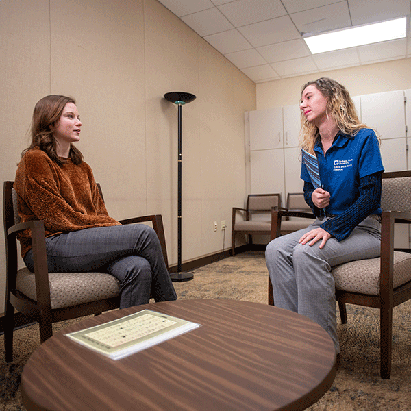 Two female students sitting in brown chairs in a room. On the left is a white female student with shoulder-length brown hair. She wears a brown long-sleeved sweater and grey dress pants. On the right is a white female student with curly blond hair. She wears a blue Indiana State University shirt with grey dress pants. A brown table is in front of them and other chairs and filing cabinets are in the background.