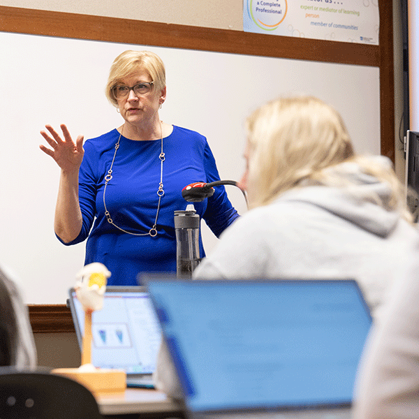 A white female professor stands at the front of a classroom. She has short blond hair, and she wears glasses, a long-sleeved blue dress, and a long gold necklace. A white female student with blond hair, wearing a white sweatshirt, sits in front of the professor. Laptops are on the desks. A whiteboard is in the background.