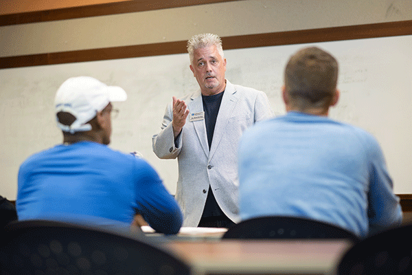 A white male professor stands at the front of a classroom. With white spiked hair, he wears a white dress suit with a dark blue/black undershirt. His right hand is raised toward two male students sitting at desks in front of him. On the left is a white male student wearing a blue long-sleeved shirt and a white baseball cap. On the right is a white male student wearing a blue long-sleeved shirt. A whiteboard is in the background.
