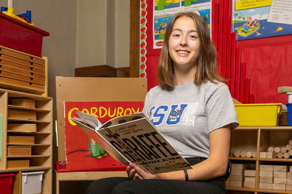 A smiling Indiana State education student sits in an elementary classroom. She is wearing a grey shirt that says ISU in blue letters and has an image of Sycamore Sam’s head inside the U. She is holding a book with the title “Giraffe Problems” that has the image of a giraffe’s neck on it. The name of the author, Jory John, and the illustrator, Lane Smith, are also visible at the top of the book. Behind the student is a red sign with the word “Corduroy” on it.