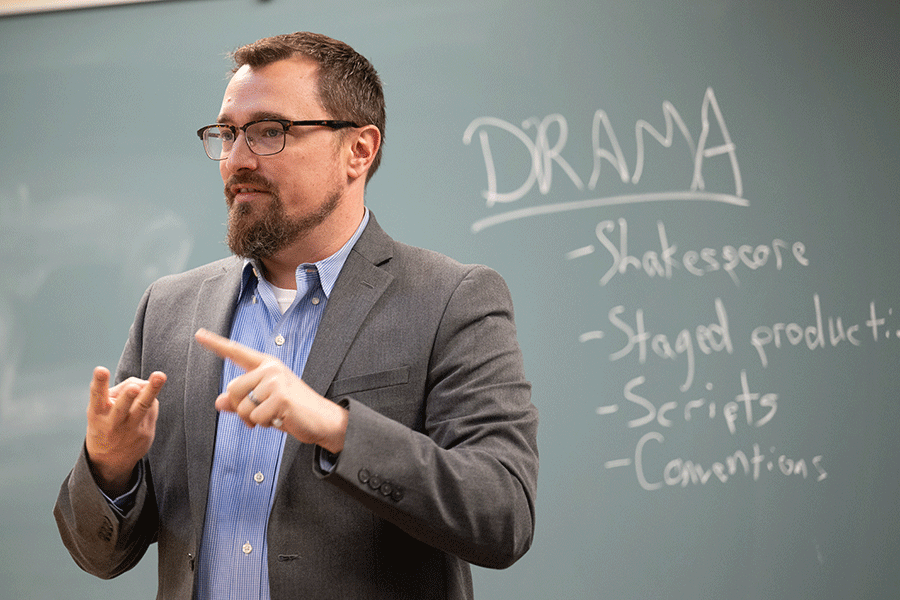 A white male professor with short, dark brown hair and glasses stands at the front of a classroom with the words Drama, Shakespeare, Staged Production, Scripts, and Conventions written on the chalkboard  behind him. He wears a grey suit jacket with a blue open-collared shirt, and his left hand is pointing to his right hand, on which three fingers are held up as if counting.