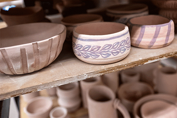 A brown shelf is visible with rows of unglazed pottery. The pottery pieces are shaped into bowls. The bowl on the left has five stripes visible from top to bottom. The bowl in the center has blue floral designs. The bowl on the right has three blue stripes slanted from top to bottom. Below this shelf is another shelf visible with clay pottery pieces.