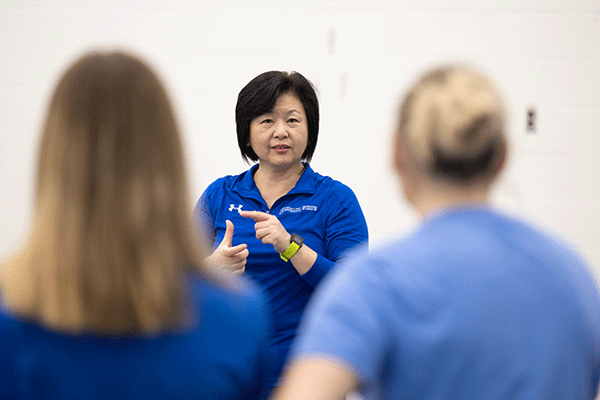 A woman of Asian heritage stands facing two women with their backs facing the camera. The woman has shoulder-length black hair, and she wears a blue Indiana State jacket and a yellow watch. The women opposite her have blond hair and are wearing blue medical scrubs, blurred in the photo.