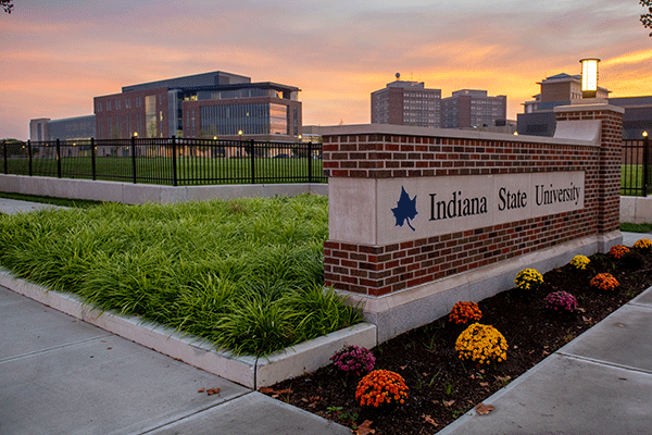 A brick stone sign reading “Indiana State University” with green grass and colorful flowers surrounding it. In the distance is the College of Health and Human Services building, a brick building with many glass windows. Other buildings are in the background.