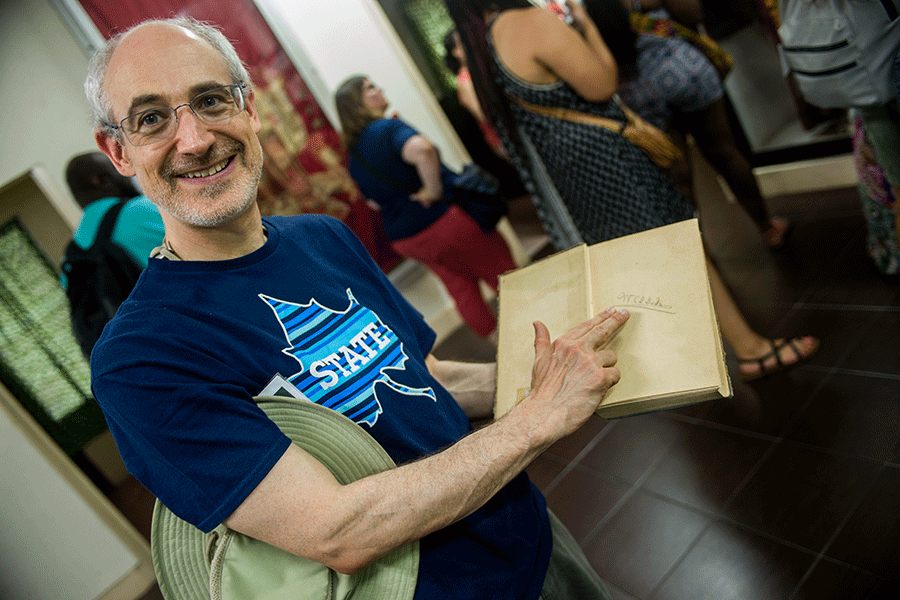 Older male adult in navy Indiana State University t-shirt smiles while pointing to a signature written in an old book.