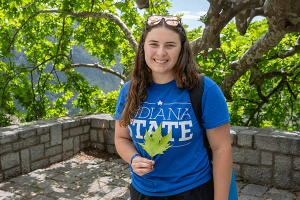 A female student with long brown hair poses outside with sunglasses on top of her head. She wears a blue Indiana State T-shirt and a backpack. She holds a green leaf. Behind her is a row of brick stones and tree branches.