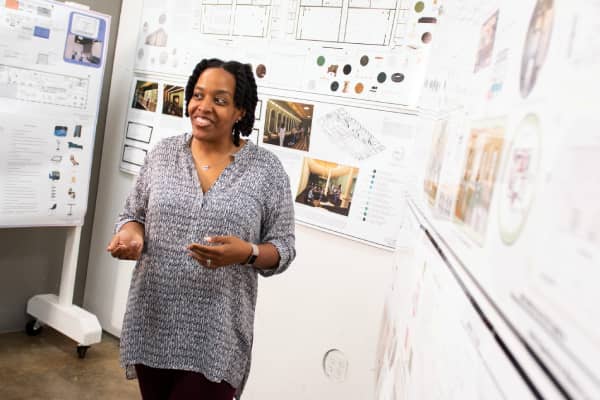 Azizi Arrington-Slocum, a Black female professor of interior architecture design, stands before three walls of interior architecture blueprints and plans. She has shoulder-length black hair, and she wears a grey shirt and black pants.