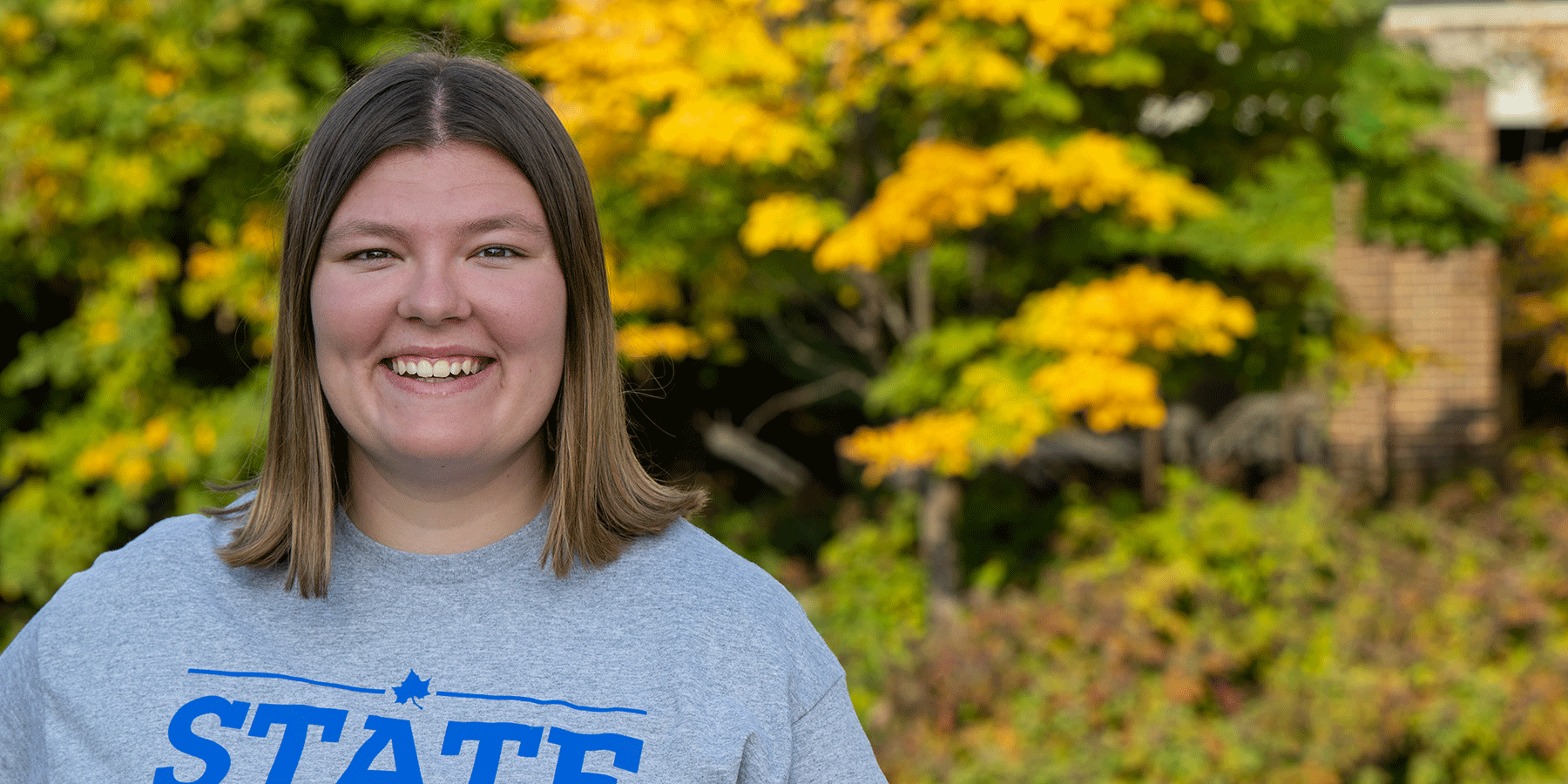A white female student with shoulder-length brown hair poses outside, smiling. She wears a grey T-shirt with STATE in blue lettering on the front. Green trees with a variety of light yellow/orange leaves are blurred in the background.