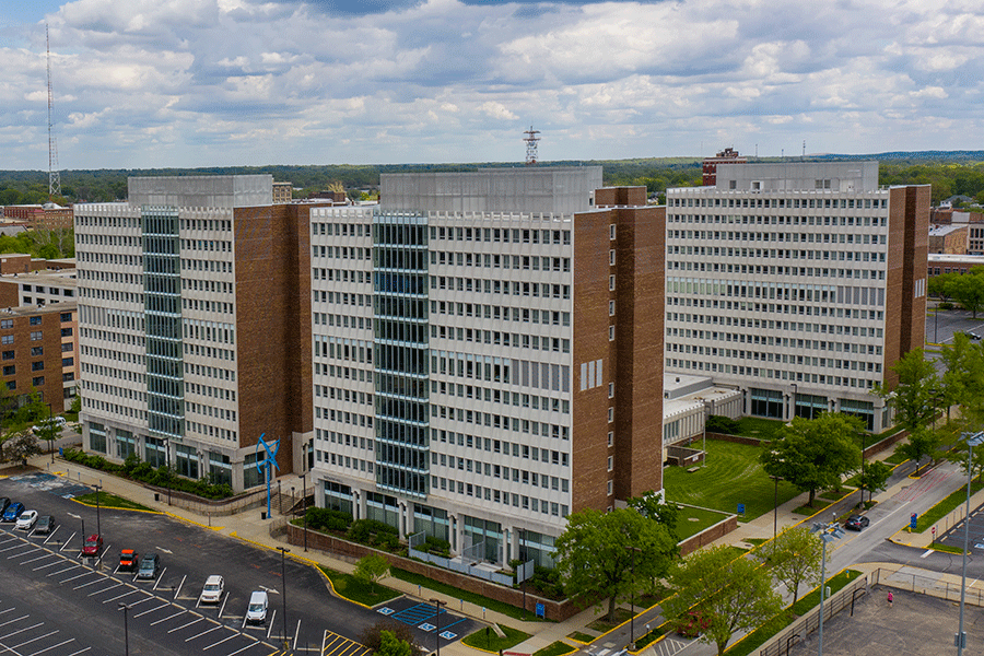An exterior view of three identical tall brick buildings with rows of glass windows. A parking lot with parked cars is in the bottom left corner of the photo. Grass and green trees line the sidewalks in the bottom right corner of the photo. There is a view of the sky with clouds.