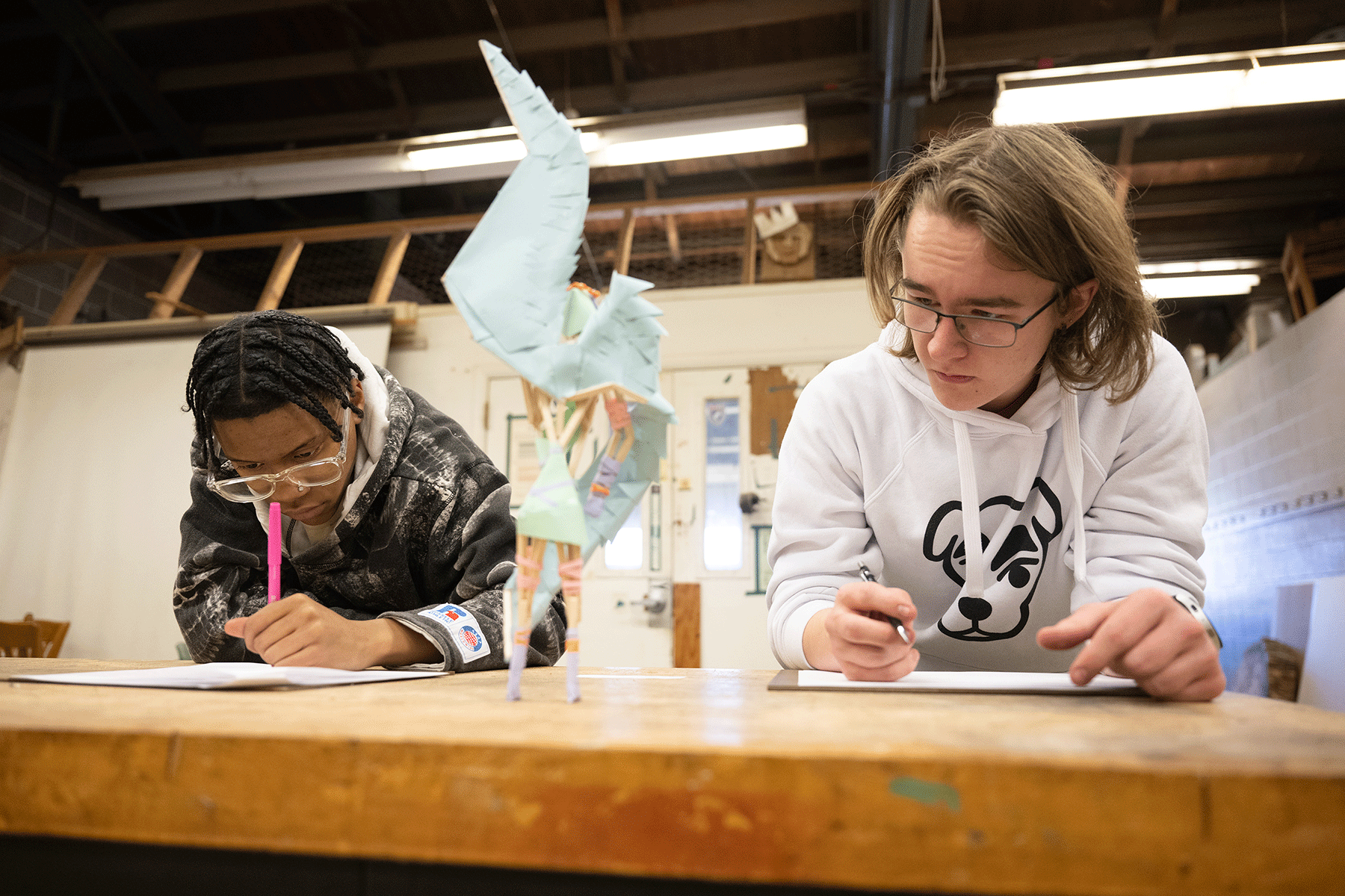 Two students seated in a workshop observe a sculpture that is on a table while writing their observations on paper.