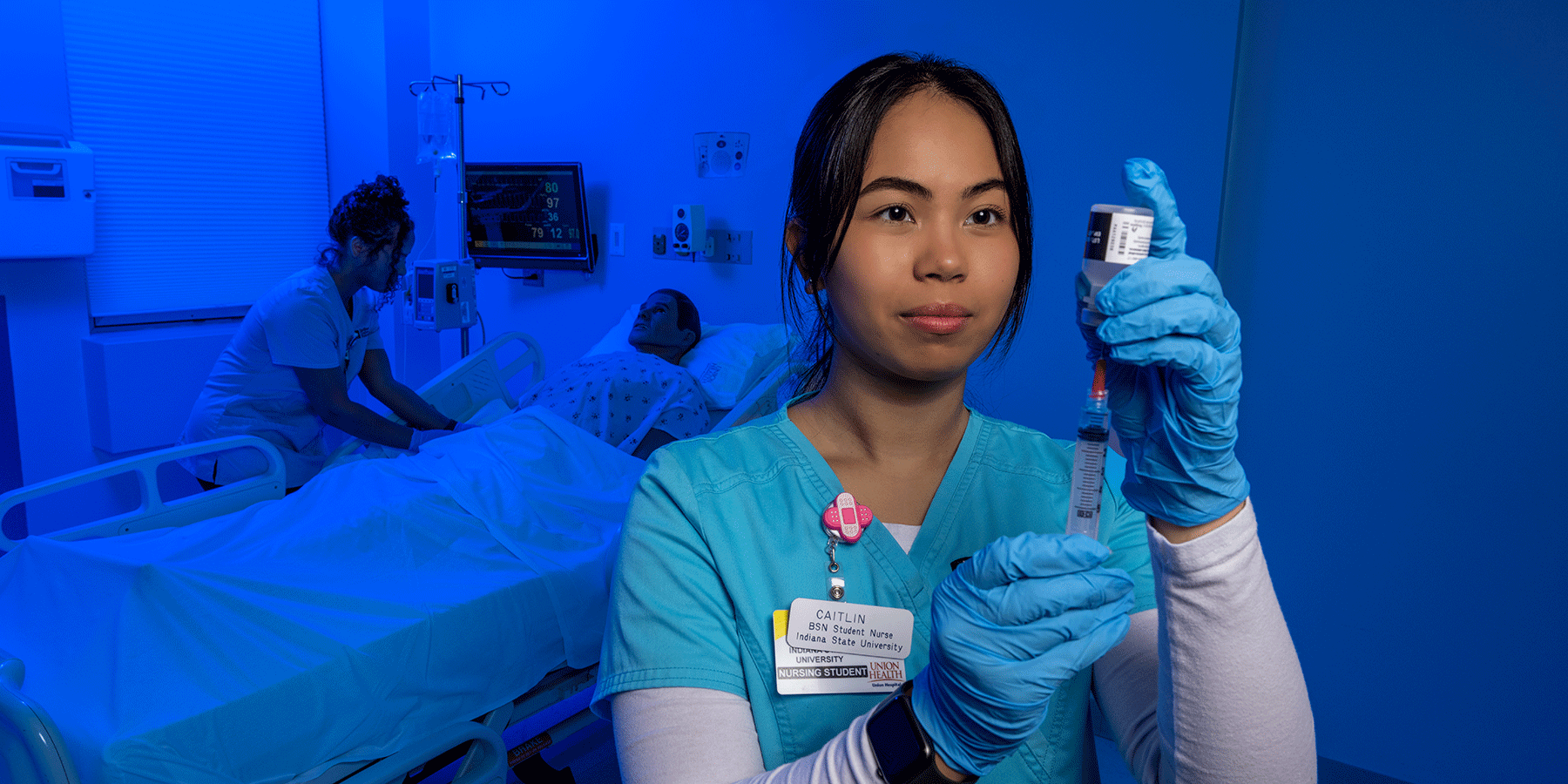 Student wearing scrubs and medical gloves uses a syringe to extract medication from a vial while another student tends to a dummy in a hospital bed in the background