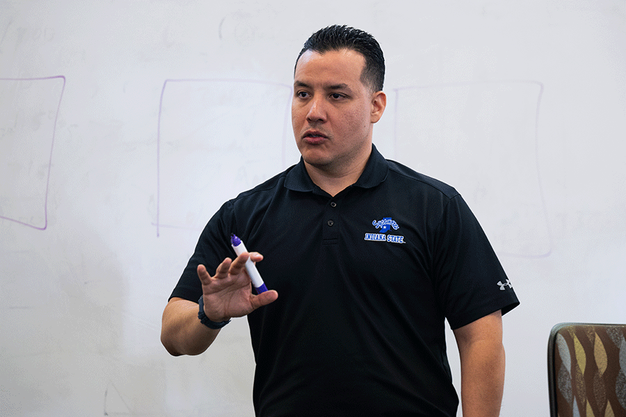 A male wearing a black polo holds a marker and stands in front of a white board while speaking