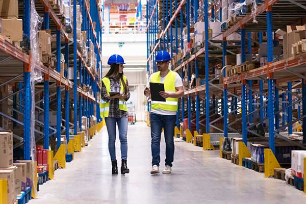Two individuals in yellow safety vests and hard hats walk through a warehouse holding clipboards
