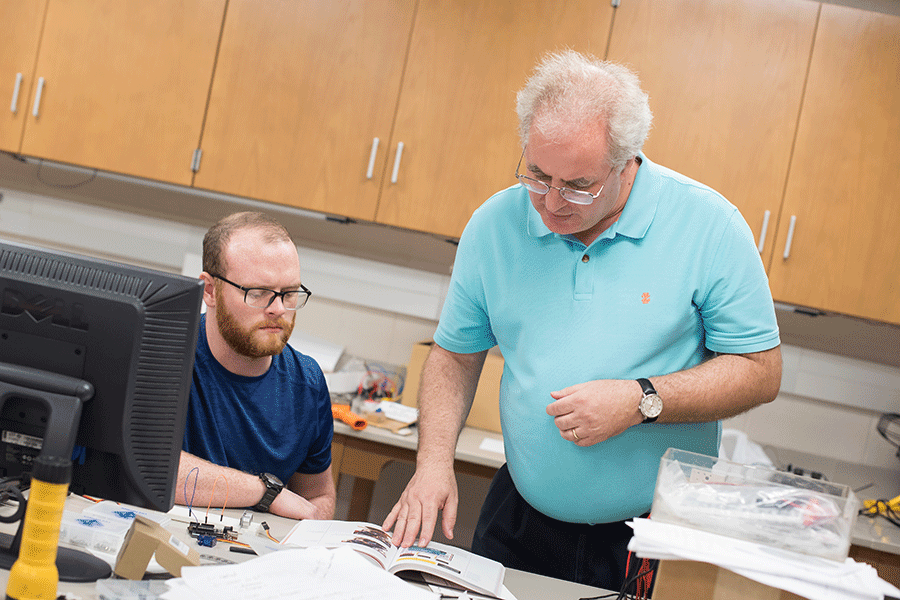 Two men are in a classroom. On the left is a man with short brown hair, with a matching beard and mustache, and glasses. He sits at a table, wearing a dark blue T-shirt. On the right is a man with short white hair and glasses. He is standing and wearing a light green polo shirt. On the table in front of them is an open textbook and other papers, with a desktop computer to the left. Brown cabinets are behind them.