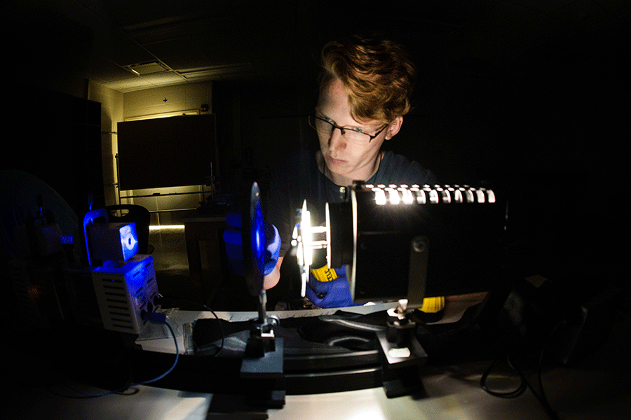 A male student with short red hair and glasses sits in a dark room. He is looking at physics lab equipment that emits an illuminating blue light.